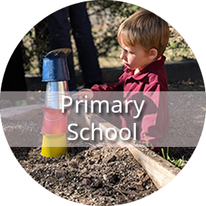 The text "Primary School" is displayed in front of a preschool child in a long sleeve burgundy polo shirt sits on the ground outside. He has stacked four buckets upside down on top of each other to create a tower with yellow on the bottom followed by red, silver and blue on the top.